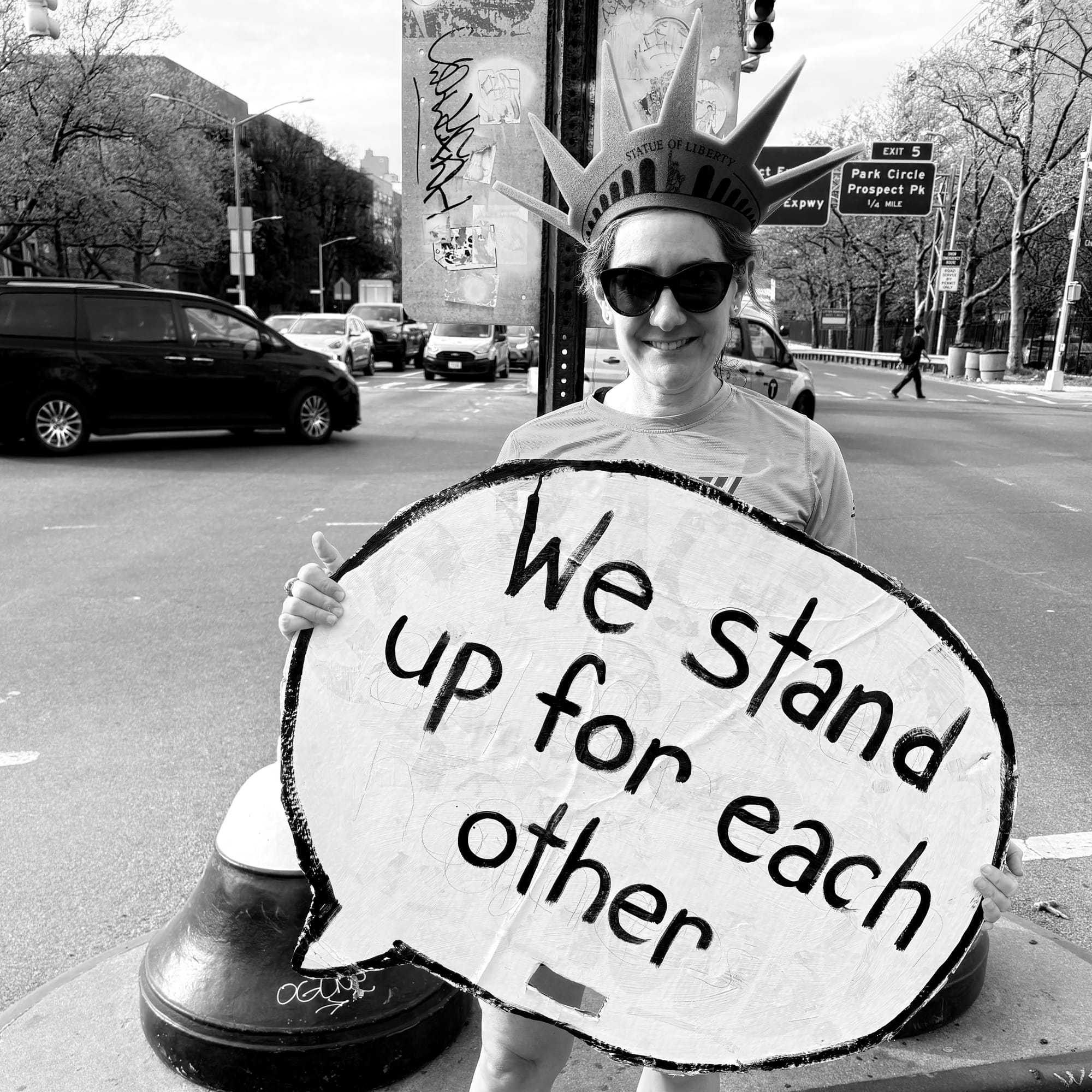 a smiling woman wearing a Lady Liberty crown, standing on the median of Ocean Parkway in Brooklyn, and holding a sign that says We stand up for each other